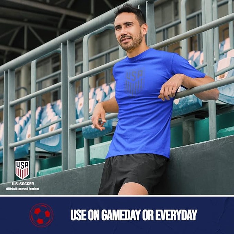 May include: A man wearing a royal blue t-shirt with a gray USA graphic and black shorts. He is standing in a stadium with bleachers in the background. The text "USA" is visible in the lower left corner of the image. The text "U.S. SOCCER Official Licensed Product" is also visible. The text "USE ON GAMEDAY OR EVERYDAY" is visible in the lower right corner of the image.