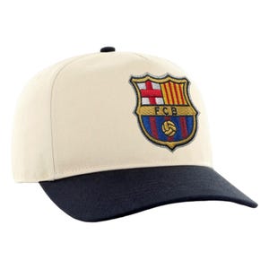 May include: A cream-colored baseball cap with a navy blue brim. The cap features an embroidered emblem with the letters "FCB" and a shield design in red, yellow, blue, and gold. The cap is a classic, adjustable style.