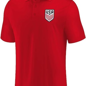 May include: A bright red short-sleeved polo shirt featuring a collar with two buttons. On the left chest, there is a shield-shaped emblem with the text USA and red and white vertical stripes. The inner neck label also displays the USA shield logo.