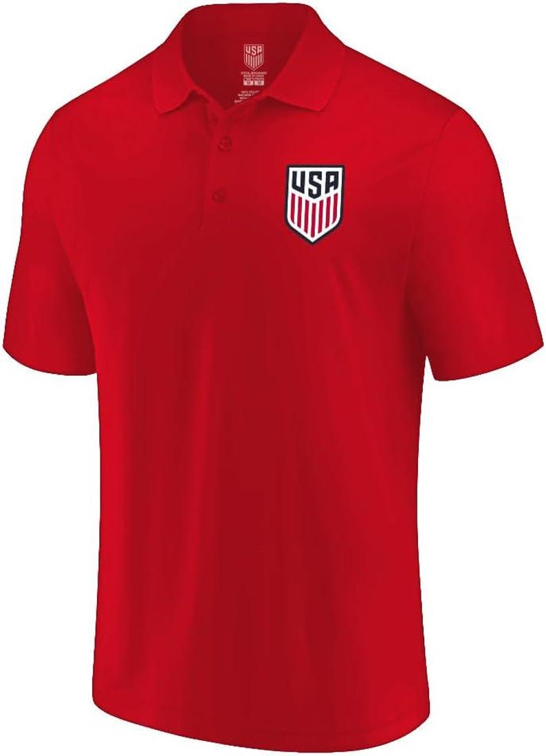May include: A bright red short-sleeved polo shirt featuring a collar with two buttons. On the left chest, there is a shield-shaped emblem with the text USA and red and white vertical stripes. The inner neck label also displays the USA shield logo.