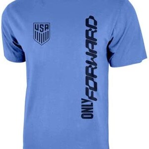 Op de afbeelding: Een lichtblauw t-shirt met een donkerblauwe afbeelding van het USA-logo en de tekst "ONLY FORWARD" die verticaal aan de rechterkant is gedrukt.