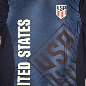 May include: A navy blue and red soccer jersey with the words "United States" and "USA" printed on it. The jersey has a striped pattern and a red collar.