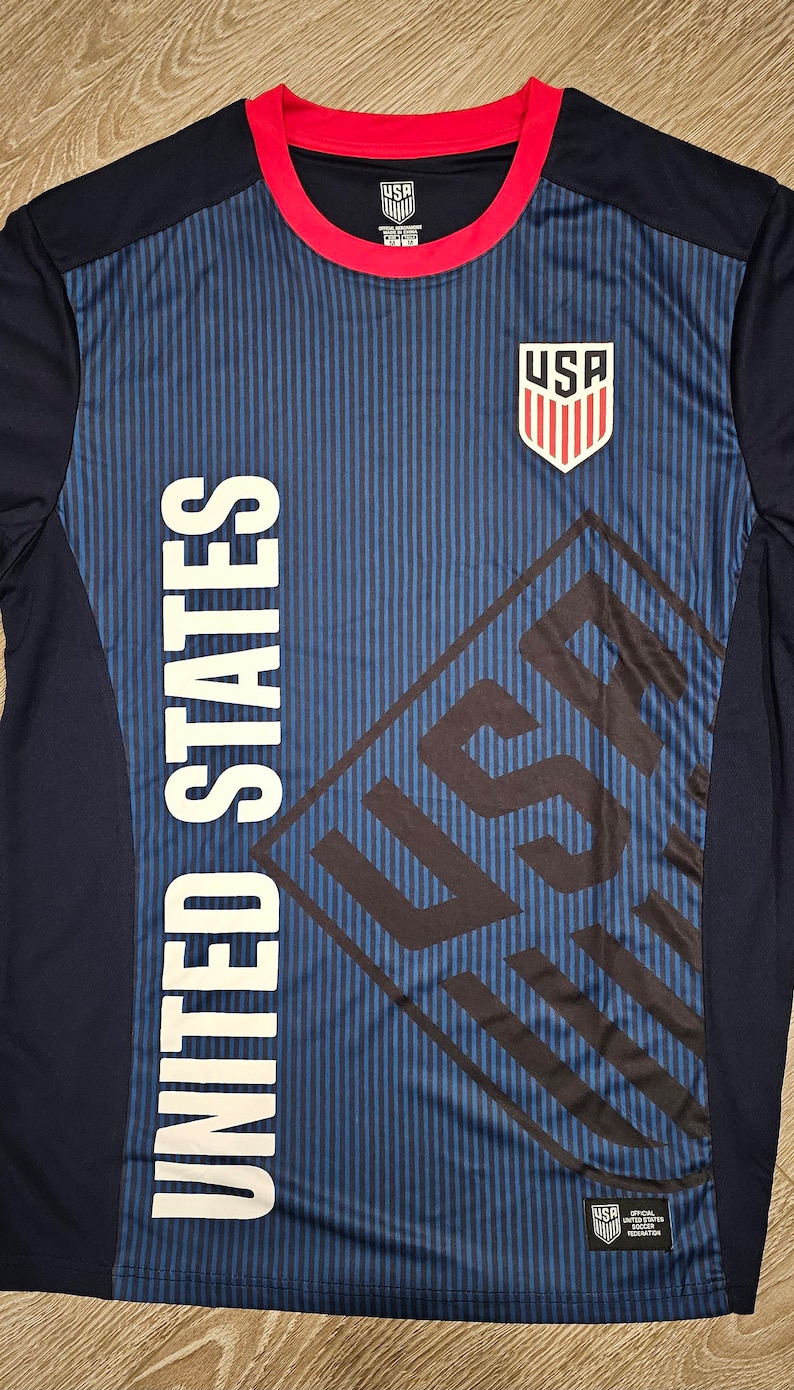 May include: A navy blue and red soccer jersey with the words "United States" and "USA" printed on it. The jersey has a striped pattern and a red collar.