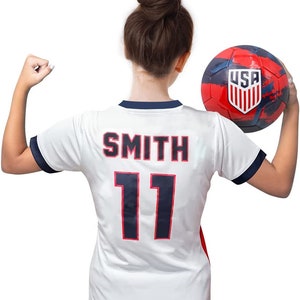 May include: A white soccer jersey with the name "SMITH" and the number "11" in red and navy blue. The jersey has navy blue trim. A red, white, and blue soccer ball with the USA logo is held in the hand.