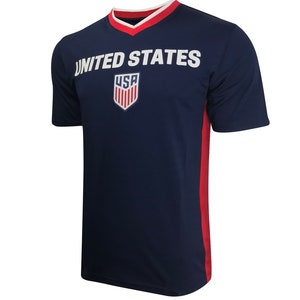 Pu&ograve; includere: Una maglietta blu navy a maniche corte con un collo a V rosso e bianco. La maglietta ha una scritta bianca "UNITED STATES" sul davanti e uno scudo rosso, bianco e blu con la scritta "USA" all'interno.