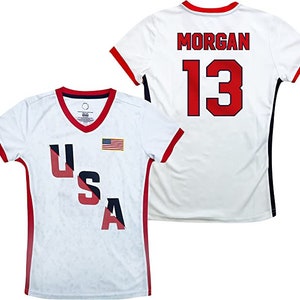 May include: White soccer jersey with red and blue USA lettering and an American flag on the front. The back of the jersey has the name "MORGAN" and the number "13" in red.