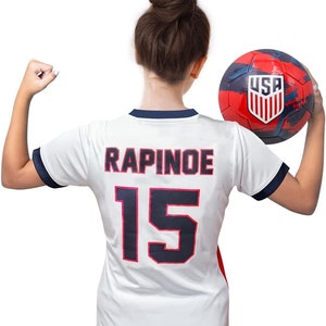 May include: White soccer jersey with the name "RAPINOE" and the number "15" on the back. The jersey has a navy blue collar and sleeves. A person is holding a red, white, and blue soccer ball with the word "USA" on it.