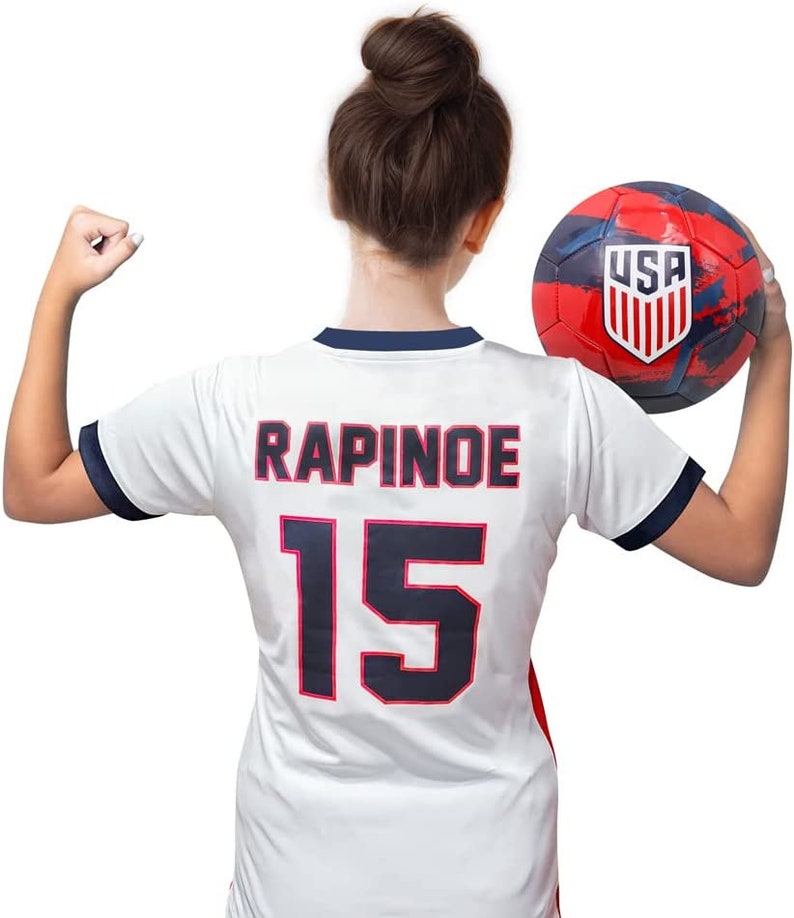 May include: White soccer jersey with the name "RAPINOE" and the number "15" on the back. The jersey has a navy blue collar and sleeves. A person is holding a red, white, and blue soccer ball with the word "USA" on it.