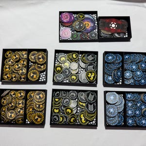 May include: A collection of game tokens in black trays. The tokens are various colors, including gold, blue, purple, and gray. Some tokens have symbols and designs. The trays are arranged on a white surface.