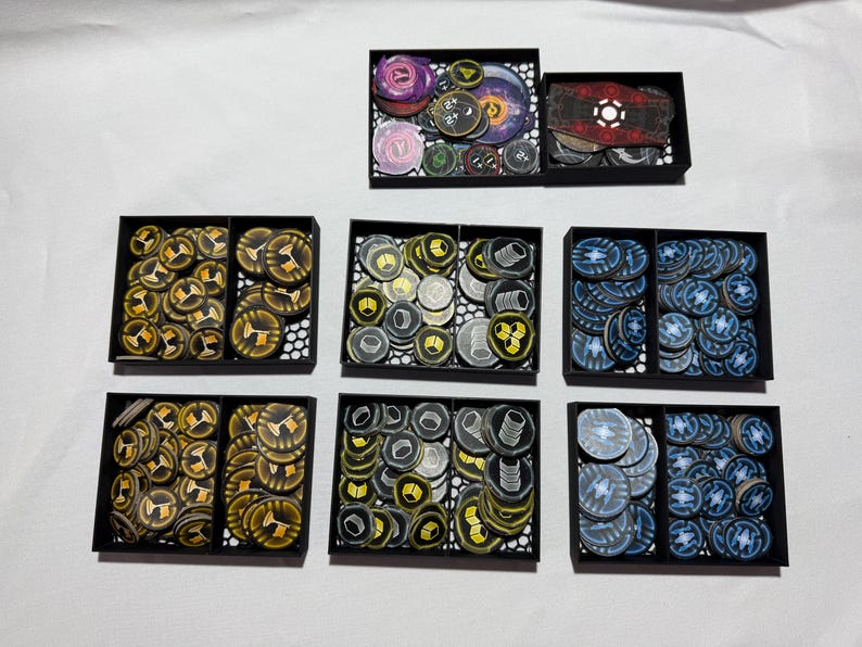 May include: A collection of game tokens in black trays. The tokens are various colors, including gold, blue, purple, and gray. Some tokens have symbols and designs. The trays are arranged on a white surface.