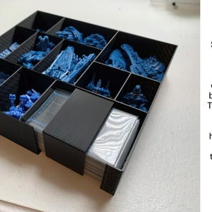 May include: A black storage box with multiple compartments, filled with blue miniature figures. A small black rectangle is partially covering a stack of cards. Text on the side explains the use of the rectangle.
