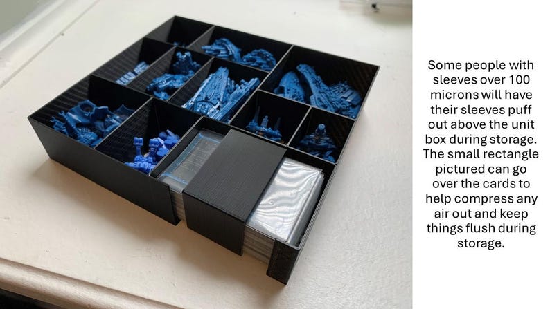 May include: A black storage box with multiple compartments, filled with blue miniature figures. A small black rectangle is partially covering a stack of cards. Text on the side explains the use of the rectangle.