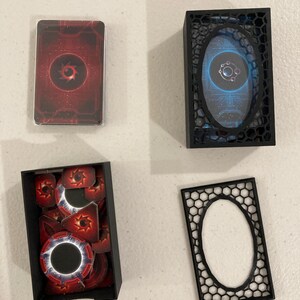 May include: A collection of game components, including a deck of cards in a plastic sleeve, tokens, and storage boxes. The cards and tokens feature a red and black design with a central circular emblem. The boxes are black.