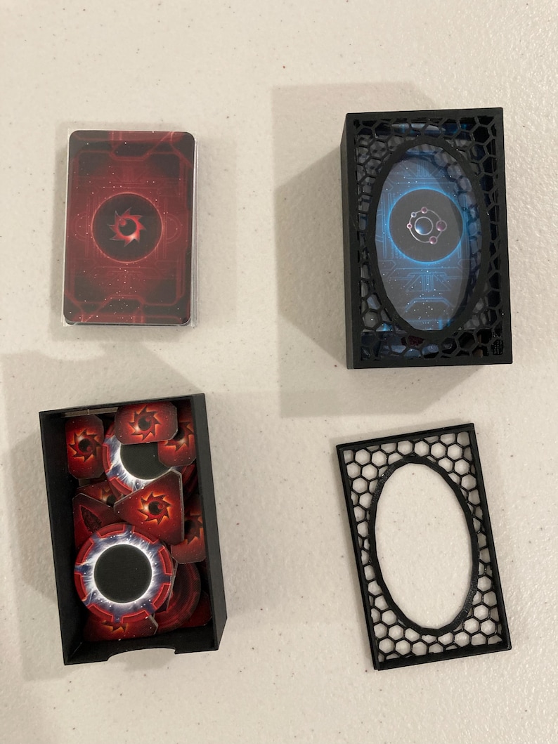 May include: A collection of game components, including a deck of cards in a plastic sleeve, tokens, and storage boxes. The cards and tokens feature a red and black design with a central circular emblem. The boxes are black.
