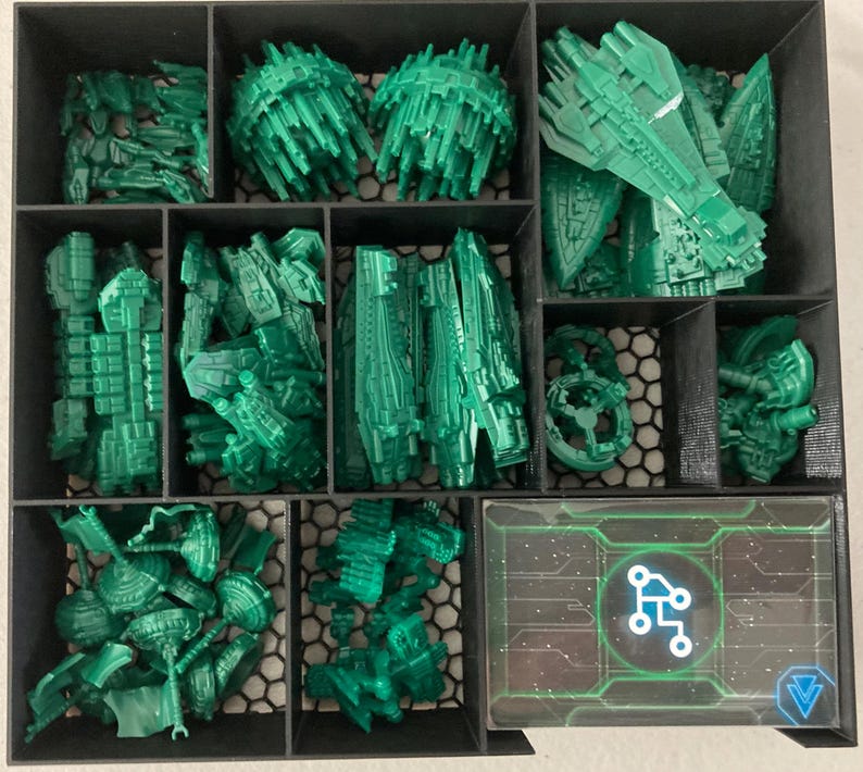 May include: A black organizer filled with various green plastic game pieces, including spaceship models and other futuristic components. A card with a circuit board design is visible in the bottom right corner.