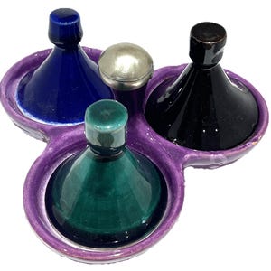 May include: A purple ceramic serving tray with three circular compartments, each holding a conical lid. The lids are glossy blue, black, and green. A small, round, metallic silver-toned object sits in the center between the three compartments.