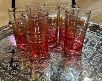 May include: Six small, clear glass shot glasses with gold trim and a pink tint are arranged on a silver tray with an ornate design.