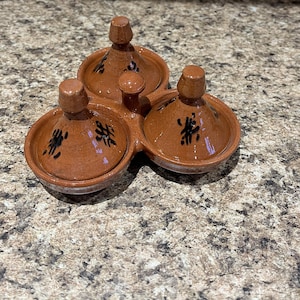 May include: A terracotta-colored ceramic condiment server with three connected bowls, each with a lid and a small handle. Black abstract designs decorate the lids. The server is designed for serving sauces or spices.