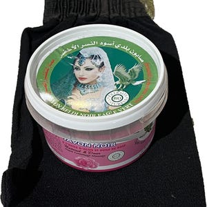 May include: A black and green jar of black soap with a woman's face on the label. The jar is labeled "Savon Noir" and "A l'huile d'olive et huile de rose." The jar is sitting on a black washcloth.