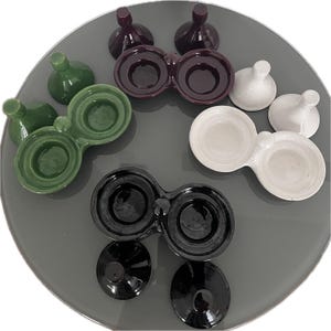 May include: A selection of ceramic condiment dishes and matching small, round-topped containers in various colours. The colours include dark green, light green, white, black, and a deep burgundy. The dishes have two connected bowls.