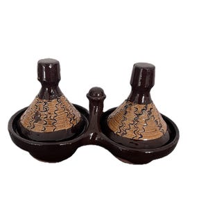 May include: A double-chambered ceramic condiment server featuring two conical lids with dark brown glaze and tan sections decorated with black wavy vertical lines. The base is a single connected piece with a central handle between the two bowls.