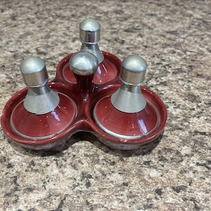 May include: A red ceramic condiment server with three sections, each topped with a silver-colored lid and a spring-like neck. The server is designed with a clover-like shape.