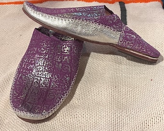 Op de afbeelding: Een paar paarse leren slippers met zilveren accenten. De slippers hebben een spitse neus en een platte zool.