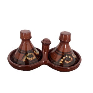 May include: A brown glazed ceramic double condiment server featuring two conical lids with a central handle. Each base holds a small tajine-shaped vessel decorated with dark brown horizontal bands and small yellow painted circular accents.