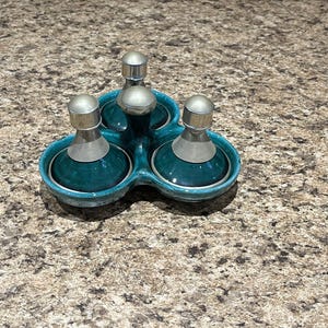 May include: A teal-colored condiment set with three glass bottles, each topped with a silver lid and a ribbed silver neck. The bottles sit in a matching teal ceramic holder with a clover-like shape. The set is on a speckled brown surface.