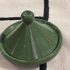May include: A green ceramic tagine pot with a conical lid and a rounded handle. The pot is a solid, glossy green colour. The tagine sits on a textured, neutral-coloured surface.