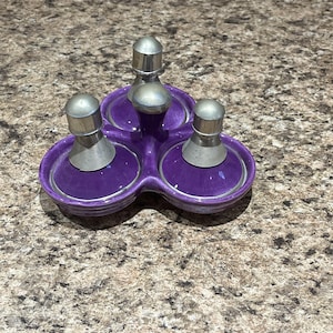 May include: A purple ceramic condiment set with three sections, each holding a silver-topped shaker. The set has a silver trim and is designed to hold salt, pepper, and other spices. The set is on a speckled brown surface.