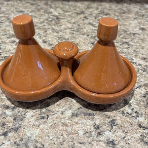 May include: A terracotta-colored ceramic salt and pepper set. The set features two conical-shaped salt and pepper shakers with rounded tops, and a small handle in the middle. The shakers sit on a connected, oval-shaped base.