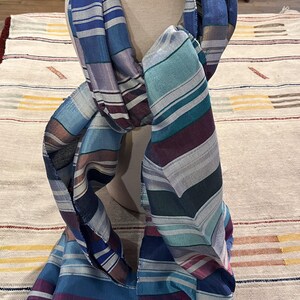 May include: A striped scarf in shades of blue, purple, teal, and gray is draped over a mannequin. The scarf has a fringed edge and a variety of stripe widths. The scarf is made of a soft, lightweight material.