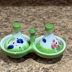 May include: A ceramic salt and pepper shaker set. The set features two small, bulbous bottles with green lids, set in a connected green tray. The bottles are decorated with blue and red floral designs on a white background.