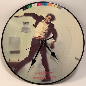 May include: A black vinyl record clock featuring a photo of David Bowie from the album "Fantastic Voyage". The clock face is white with a grid pattern and the album title is printed in black text. The record label is RCA and the album number is BOWP 109 (BOW 2).