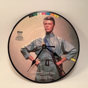 May include: A black vinyl record clock featuring a photo of David Bowie on the album cover for "Fashion". The album title is printed in rainbow colors. The clock face is black with white hands. The album cover features a photo of David Bowie wearing a blue shirt and a black belt. The album title "A New Career in a New Town" is printed on the bottom of the record.
