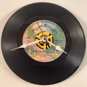 May include: A black vinyl record clock featuring the song "Shake, Rattle and Roll" by Bill Haley and His Comets. The clock has white hands and a yellow center, with the text "Burbank, Home of Warner Bros. Records."
