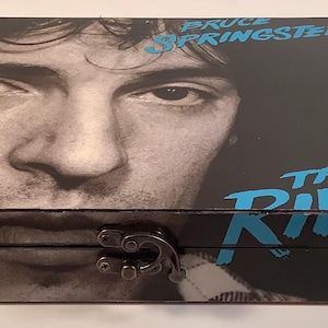 May include: A black wooden box with a hinged lid. The lid features a black and white photo of Bruce Springsteen, with the text "Bruce Springsteen" and "The River" in blue lettering.