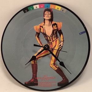 May include: A clock featuring a record from the album "Drive-In Saturday" by David Bowie. The clock face features a color image of David Bowie in a striped jumpsuit. The clock hands are black. The record is labeled with the album title, "Drive-In Saturday", and the side, "Side A".