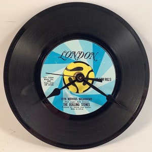 May include: A black vinyl record clock with a light blue and yellow center. The clock face features the text "LONDON" and "19th NERVOUS BREAKDOWN" by The Rolling Stones. The clock hands are black.