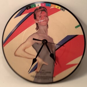May include: A vinyl record clock featuring a photo of David Bowie from the album "Fashion" with a red, white, and blue design. The clock hands are black and the clock face is white.