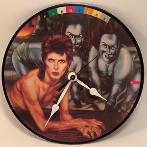 May include: A clock with a black frame and a white face. The clock face features a colorful image of David Bowie from his album "Rebel Rebel". The album cover features a colorful image of David Bowie with two women. The album title "Rebel Rebel" is printed on the clock face.
