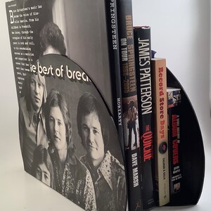 May include: A set of black record-shaped bookends with a black and white photo of a band on the front. The bookends are holding up a collection of books about Bruce Springsteen, including "Bruce Springsteen on Tour 1968-2005" by Dave Marsh and "The Quickie" by James Patterson.