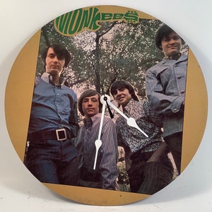 May include: A vintage clock featuring a gold background with a picture of the band The Monkees. The clock has black hands and a white face. The band members are dressed in 1960s clothing and are standing in front of a green leafy background.