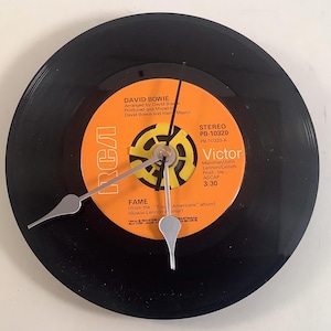 May include: A black vinyl record clock featuring a vibrant orange and yellow center label with the text "DAVID BOWIE" and "FAME". The clock hands are silver, and the clock is mounted on a white surface.