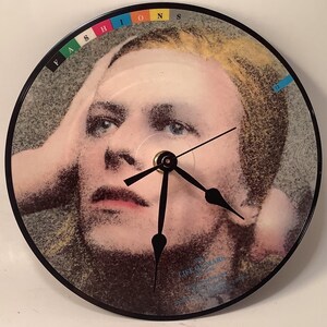 May include: A black vinyl record clock featuring a close-up image of David Bowie's face. The clock face is a black circle with white numbers and black hands. The record label is visible with the text "SP 125 LIKE A PRAYER" and "David Bowie - 1983".