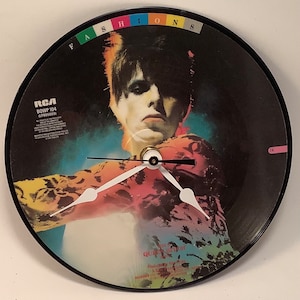 May include: A black vinyl record clock featuring a colorful image of David Bowie from the album "Fashion" by David Bowie. The record label reads "RCA BOWP 104 (LPB05009)".