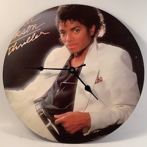 May include: A white vinyl record clock featuring a black and white image of Michael Jackson on the cover of his album "Thriller". The clock hands are black and the album title is written in gold lettering.
