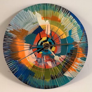 May include: A round wall clock made from a vinyl record, featuring a colorful abstract design. The clock face has a vibrant mix of orange, blue, and green paint splatters radiating from the center. The clock hands are a metallic gold color.