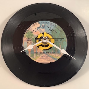 May include: A black vinyl record clock with white clock hands. The record label features text including "Rock Around the Clock" and "Bill Haley And His Comets". The clock has a yellow and black center.